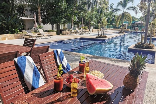 Resort pool area with wooden lounge chairs, refreshments on a table, palm trees, and sunbathing guests in the background
