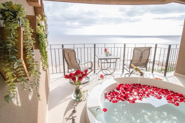 Oceanfront bathroom with soaking tub filled with red rose petals, hanging plants, and balcony seating overlooking calm sea.